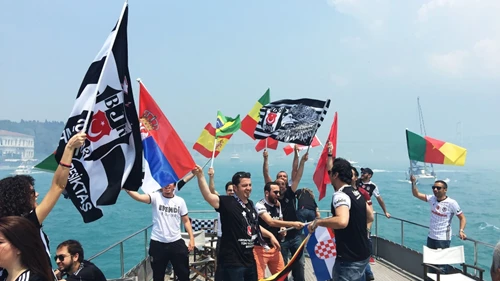 Beşiktaş-Fans feiern mit Vereinsfahnen und internationalen Flaggen auf einem Boot auf dem Bosporus vor der Kulisse Istanbuls.